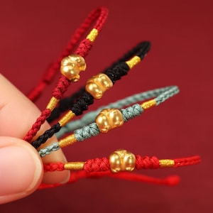 Op de afbeelding: Close-up van verschillende gevlochten armbanden in rood, zwart en blauwgroen. Elke armband heeft een goudkleurige bedel en gouden accenten. De armbanden worden getoond tegen een rode achtergrond.