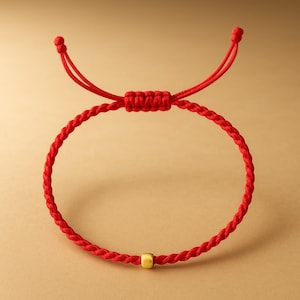 Può includere: Un braccialetto rosso intrecciato con chiusura scorrevole regolabile. Il braccialetto presenta una piccola perla quadrata color oro. Lo sfondo è di colore beige neutro.