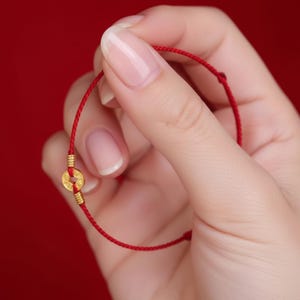 May include: Red string bracelet with a gold coin charm. The bracelet features a thin, vibrant red cord and a small, circular gold coin pendant. The coin has a hole in the center and is attached with gold accents. The background is a solid red.