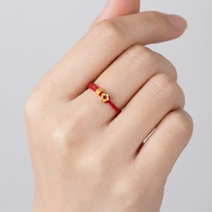 May include: A red string ring with a small gold flower and a gold bead. The ring is worn on a finger. The flower has a small red gem in the center. The ring is a delicate piece of jewelry.