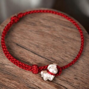 May include: A red braided bracelet with a silver charm shaped like a small animal. The bracelet is knotted at the ends and around the charm. The charm is a light silver color and has a detailed design.