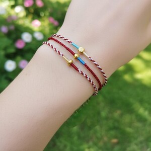 Puede incluir: Primer plano de tres pulseras delicadas. Cada pulsera presenta un fino cord&oacute;n trenzado en rojo y blanco o rojo liso. Los detalles incluyen cuentas doradas y peque&ntilde;as secciones azules. Las pulseras se llevan en una mu&ntilde;eca, con un fondo verde borroso.