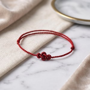 May include: A red string bracelet with a knotted design. The bracelet is made of two strands of red cord, with a knot in the center. The bracelet is lying on a white marble surface.