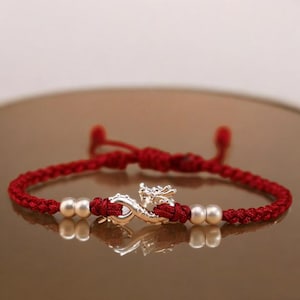 May include: Red braided bracelet with a silver dragon charm and two silver beads. The bracelet is tied with a sliding knot closure. The dragon charm is detailed and the bracelet is displayed on a reflective surface.