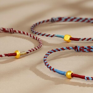 Puede incluir: Primer plano de tres pulseras trenzadas. Las pulseras presentan una combinaci&oacute;n de hilos rojos, blancos y azules. Cada pulsera tiene una cuenta dorada. Las pulseras est&aacute;n sobre una superficie de color neutro.