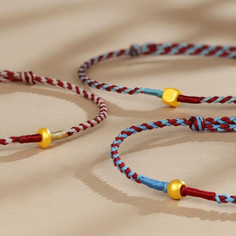Puede incluir: Primer plano de tres pulseras trenzadas. Las pulseras presentan una combinaci&oacute;n de hilos rojos, blancos y azules. Cada pulsera tiene una cuenta dorada. Las pulseras est&aacute;n sobre una superficie de color neutro.