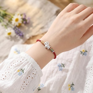 Op de afbeelding: Rode koordarmband met een zilveren ram-bedel. De armband heeft een geweven gedeelte met kleurrijke strepen. De ram-bedel heeft gedetailleerde hoorns en gelaatstrekken. De armband wordt om de pols gedragen.