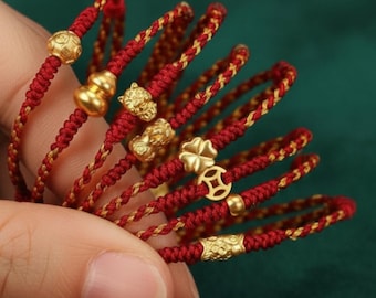 Puede incluir: Primer plano de varias pulseras trenzadas rojas y doradas con abalorios dorados. Los abalorios incluyen una moneda, una calabaza, un tigre, un trébol de cuatro hojas y un diseño geométrico. Las pulseras se sostienen sobre un fondo verde oscuro.