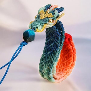 May include: A handcrafted macrame dragon ornament in shades of blue, green, and orange. The dragon's head features gold accents and small golden beads. A blue gemstone hangs from a blue cord, adding a touch of elegance.
