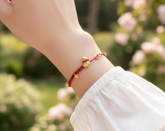 Puede incluir: Una pulsera de hilo rojo con abalorios dorados. La pulsera presenta un pequeño dije de oso, dijes de estrella y cuentas redondas. La pulsera se usa en una muñeca, con una camisa de manga blanca en el fondo.