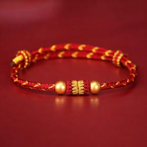 May include: A red and gold braided bracelet with gold detailing. The bracelet has two gold beads and a central gold and red decorative section. The background is a solid red.