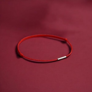 May include: A minimalist red string bracelet with a small, silver-colored cylindrical accent. The bracelet is set against a deep red background, creating a simple yet striking visual.