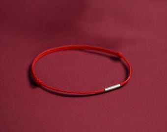 Puede incluir: Una pulsera minimalista de hilo rojo con un pequeño acento cilíndrico plateado. La pulsera se encuentra sobre un fondo rojo oscuro, creando una imagen sencilla pero llamativa.