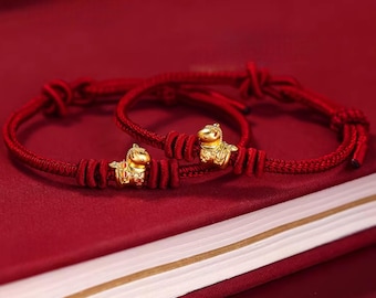 Puede incluir: Dos pulseras de cuerda roja con dijes dorados en forma de caballo. Las pulseras anudadas descansan sobre una superficie roja. Los dijes de caballo son detallados, agregando elegancia. Estas pulseras son un símbolo de buena suerte.