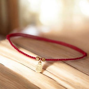 May include: A red cord bracelet featuring a gold-coloured charm. The charm has Chinese characters. The bracelet includes two small gold-coloured beads. The bracelet rests on a wooden surface.