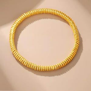 Könnte beinhalten: Ein goldfarbenes Armband mit einem spiralförmigen Design. Das Armband ist kreisförmig und scheint aus Metall zu bestehen. Das Armband hat eine leuchtend goldene Farbe und ist vor einem neutralen Hintergrund platziert.