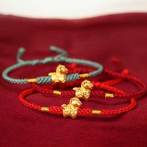 May include: Three gold charm bracelets with a braided cord design. Each bracelet has a small gold animal charm and gold accents. The cords are red and teal, set against a red background.