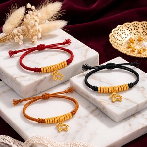 May include: Three adjustable cord bracelets in red, black, and brown, each featuring a row of small gold-toned cylindrical beads and a central gold-toned charm shaped like a stylised ram or sheep, with knotted closures and frayed ends.