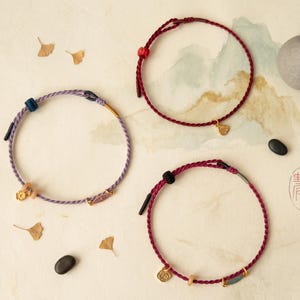 May include: Three circular bracelets made of twisted cord in shades of purple, dark red, and magenta. Each bracelet features small metallic charms, including floral and geometric shapes, along with adjustable sliding knots and metal accents.