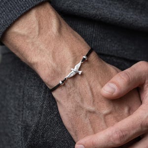 May include: A black cord bracelet featuring a central silver-toned bird charm in flight, flanked by two small silver-toned spherical beads. The bracelet is secured with a simple knot design, showcasing a clean and minimalist aesthetic on the wrist.