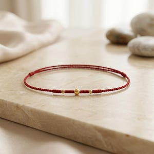 Puede incluir: Una pulsera de cordón rojo con tres cuentas doradas. La pulsera está colocada sobre una superficie clara, con una tela blanca y suave y piedras lisas en el fondo. La pulsera tiene un diseño sencillo y elegante.