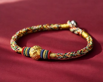 May include: A braided bracelet with a gold-coloured central bead and colourful threads. The bracelet features red, blue, green, and yellow threads, with a gold-coloured clasp. The central bead has an embossed design.