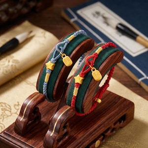 May include: Two bracelets on a wooden display. One is blue and gold, the other is red and gold. Each bracelet has a gold star and a gold charm. The bracelets are displayed on a green velvet cushion.