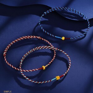 Puede incluir: Tres pulseras trenzadas en tonos rojos, azules y blancos, con peque&ntilde;os detalles amarillos y azules. Las pulseras est&aacute;n dispuestas sobre una superficie azul oscuro. La palabra "SIMPLE" est&aacute; impresa en blanco en la esquina inferior izquierda.