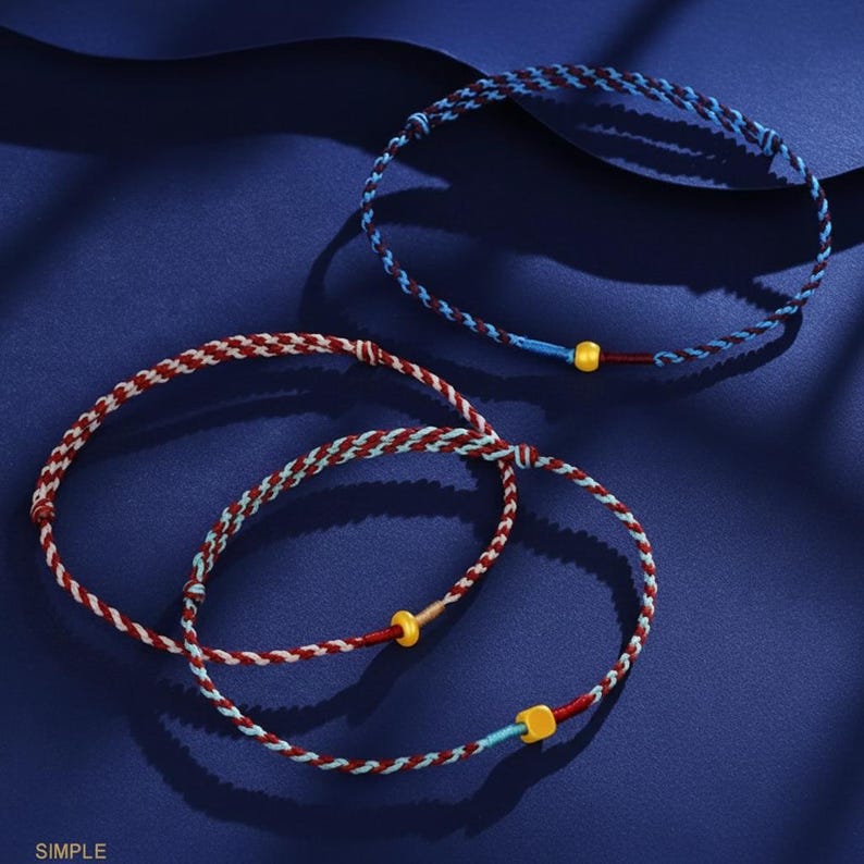 Puede incluir: Tres pulseras trenzadas en tonos rojos, azules y blancos, con peque&ntilde;os detalles amarillos y azules. Las pulseras est&aacute;n dispuestas sobre una superficie azul oscuro. La palabra "SIMPLE" est&aacute; impresa en blanco en la esquina inferior izquierda.
