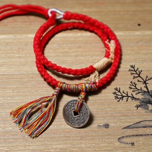 May include: A red braided cord bracelet with a decorative coin pendant and a colorful tassel. The coin has an intricate design. The bracelet is on a wooden surface.