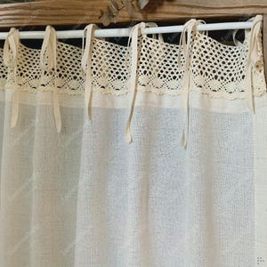 May include: Cream-colored linen curtains with a decorative lace trim at the top. The curtains are hung with fabric ties on a white rod. The fabric has a textured appearance, and the lace trim adds a delicate touch.