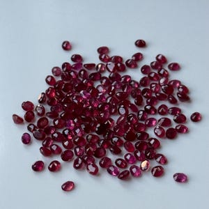 May include: A collection of small, oval-shaped, faceted rubies. The gemstones are a deep, rich red color, with a slight variation in tone. The rubies are scattered across a white surface, creating a sparkling display. Ideal for jewelry making.
