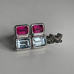 May include: A pair of stud earrings featuring a vertical arrangement of two rectangular gemstones set in silver-toned metal. The top stone is a deep pink, while the bottom stone is a pale blue. Two metal earring backs are positioned to the side.