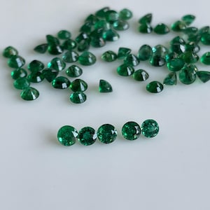 Puede incluir: Un conjunto de vibrantes gemas de esmeralda verde, tanto redondas como facetadas, dispersas sobre una superficie blanca. Las gemas varían en tamaño y forma, mostrando su brillo y color naturales.