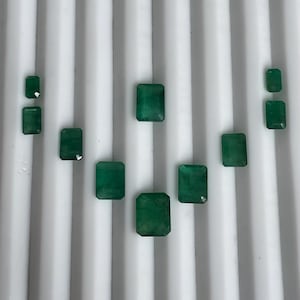 May include: A collection of nine emerald gemstones, each featuring a rectangular step-cut design. The stones vary in size and display a deep, vibrant green color with natural internal inclusions, arranged in a symmetrical pattern on a white surface.