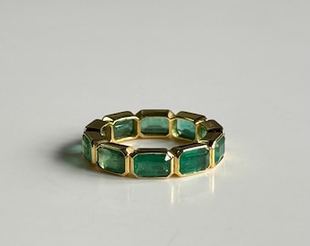 May include: A gold ring featuring emerald-cut green gemstones. The ring has a gold band and is set with rectangular emeralds. The gemstones are a vibrant green color, and the ring is displayed on a white surface.