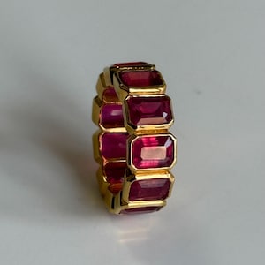 May include: A gold ring featuring a series of rectangular, deep red gemstones. The gemstones are set closely together, creating a continuous band of color. The ring has a polished, reflective surface.
