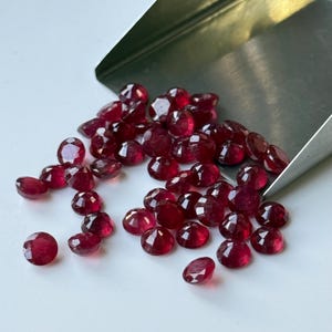 May include: A scattering of faceted, deep red gemstones, possibly rubies, on a white surface. The gems vary in size and shape, with a metallic scoop in the background. The stones have a brilliant, reflective quality.