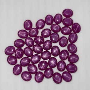 Può includere: Un gruppo di gemme ovali, viola scuro, con un effetto a stella. Le gemme lucide sono disposte su una superficie bianca. Queste gemme sono ideali per la creazione di gioielli.