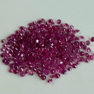 May include: A collection of small, round, faceted rubies in varying shades of deep red and pink. The gemstones are scattered across a light-colored surface, showcasing their brilliance and clarity. The gems are ideal for jewelry making.