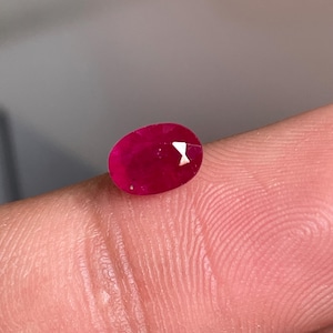 Puede incluir: Una gema de rubí de corte ovalado con un tono rojo intenso y una superficie facetada. La piedra muestra una claridad interna nítida y reflejos de luz geométricos y definidos en su parte superior pulida, exhibiendo un color carmesí saturado.