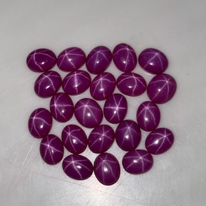 Puede incluir: Colección de gemas ovaladas de color magenta intenso. Cada gema presenta un patrón de estrella de seis rayos. Las piedras están dispuestas sobre una superficie clara, resaltando su calidad pulida y reflectante. Adecuadas para la creación de joyas.