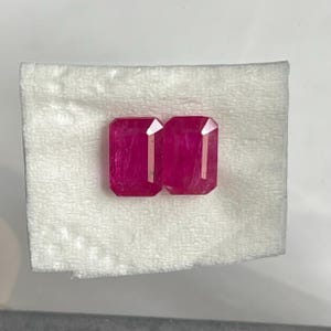 Op de afbeelding: Twee levendige, diep roze-rode edelstenen met een rechthoekige smaragdgeslepen vorm. Deze gefacetteerde stenen vertonen duidelijke interne insluitingen en scherpe, gepolijste randen die het licht reflecteren op hun oppervlakken terwijl ze naast elkaar liggen.