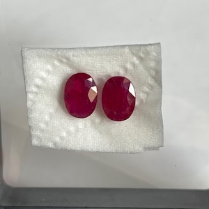 Könnte beinhalten: Zwei oval geschliffene Rubine mit einer tiefen, gesättigten roten Farbe. Die Edelsteine liegen nebeneinander und zeigen einen Brillantschliff mit mehreren reflektierenden Oberflächen, die das Licht einfangen und ihren lebendigen, intensiven roten Farbton zur Geltung bringen.