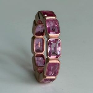 Puede incluir: Un anillo de oro rosa con una serie de piedras preciosas rosas rectangulares. Las piedras están engastadas muy juntas, creando una banda continua de color. El diseño del anillo es simple pero elegante, con un acabado pulido.