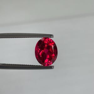 May include: A faceted, oval-shaped, deep red gemstone is held by a pair of tweezers against a white background. The gem's cut allows light to reflect, creating a brilliant display of color and sparkle. The gemstone is a vibrant shade of red.