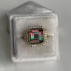 May include: A gold ring featuring a large, rectangular, multi-colored gemstone with gradient hues of pink, green, and dark tones. The center stone is surrounded by a halo of small, clear, round-cut white stones set in a gold frame.