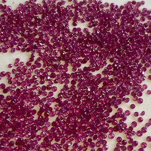 May include: A scattering of small, faceted, deep red gemstones. The gems are scattered across a light-colored surface, catching the light and displaying a brilliant shine. The gemstones appear to be rubies, with a rich, saturated color.