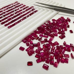 May include: A collection of radiant, rectangular-cut rubies in varying sizes. Some are neatly arranged in rows on a white tray, while others are scattered. A pair of tweezers rests nearby, suggesting gemstone handling or sorting.
