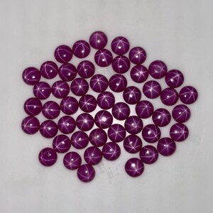 Puede incluir: Un conjunto de gemas redondas, pulidas, de color magenta oscuro, con un efecto de estrella. Cada gema tiene un patrón de estrella de seis rayos. Las gemas están dispuestas juntas sobre una superficie blanca, creando una exhibición visualmente atractiva.